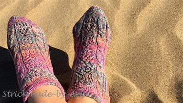 Ankle-length hand-knitted socks in variegated pink yarn showing lace stitch details on sand