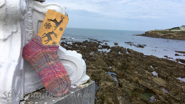 Multicolored hand-knitted ankle sock on a wooden sock blocker with deer and snowflake cutouts, set on a white ledge by a rocky coastline.