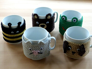 Five white mugs wearing crocheted animal cozies: bee, bear, frog, cat and dog
