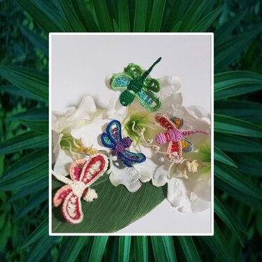 Fünf gehäkelte Libellen in verschiedenen Farben auf weißen Blüten und einem grünen Blatt.