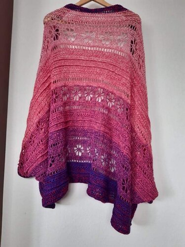 Gehäkelter Seelenwärmer, Farbverlauf Pink zu Violett, Blüten‑Lochmuster, an Holzbügel hängend.
