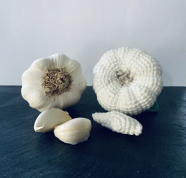 Gehäkelter Knoblauchkopf und gehäkelte Zehe neben natürlichem Knoblauch und zwei Knoblauchzehen auf dunklem Untergrund.