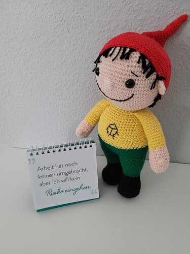 Nahaufnahme eines gehäkelten Wichtels mit roter Mütze neben einem Notizbuch mit Spruch
