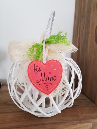 Filzherz mit 'für Mama' Stickerei an einem weißen Korb, umgeben von Netzstoff und grünen Deko-Elementen.