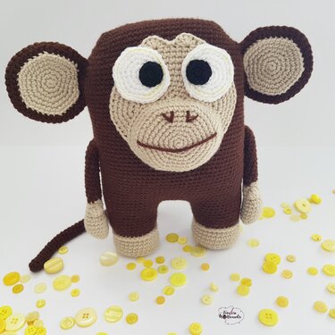 Brauner gehäkelter Amigurumi-Affe mit beigem Gesicht, auf hellem Hintergrund mit verstreuten gelben Knöpfen.