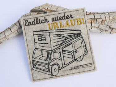 Beiger quadratischer Tassenteppich aus Filz mit gesticktem Camper und dem Schriftzug „Endlich wieder URLAUB!“.