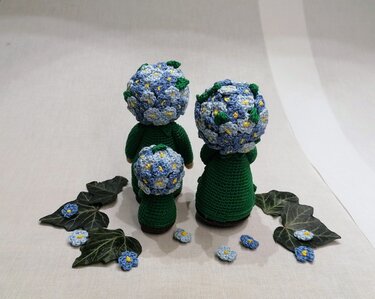 Drei gehäkelte Amigurumi-Puppen mit grünen Körpern und blauen Blütenköpfen, Rückansicht.