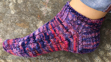 Gestrickte mehrfarbige Socke in Lila, Pink und Orange mit Zopfmuster an einem Fuß auf steiniger Oberfläche