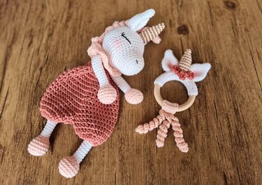 Gehäkeltes Einhorn-Schmusetuch in Weiß und Altrosa neben einem passenden Beißring mit Holzring auf Holzoberfläche