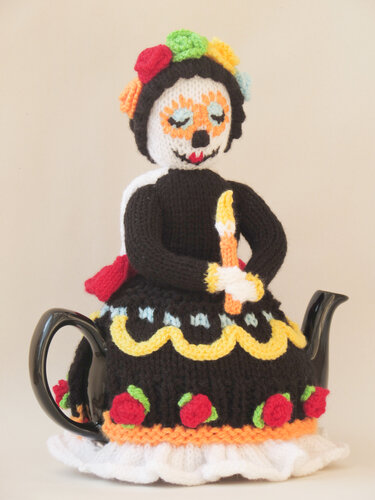 Knitted teapot cozy shaped like a sugar skull figure in a black dress with a colorful floral crown, covering a teapot.
