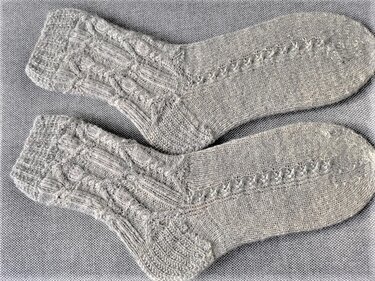 Zwei graue, handgestrickte Damensocken mit Zopf- und Lochmuster, nebeneinander auf hellgrauem Stoff.