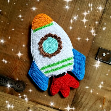 Bunte gehäkelte Rakete mit oranger Spitze, grünen Streifen, blauen Flossen und roten Flammen auf Holzuntergrund.