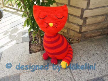 Aufrecht sitzende, rot-gestreifte Häkelkatze mit gelben Akzenten auf gepflastertem Weg neben einer Mauer.