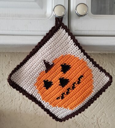 Handmade crocheted square potholder with an orange jack-o'-lantern on white, brown border, hanging from a cabinet knob.