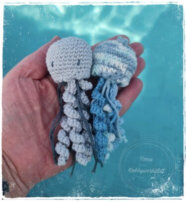 Zwei handgehäkelte Amigurumi-Quallen in einer Hand vor blauem, unscharfem Hintergrund; eine hellgrau, eine blau-weiß gestreift.