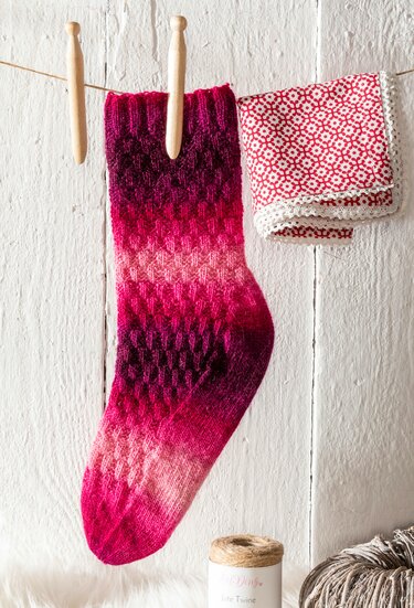 Gestrickte Damensocke in Magenta- und Pinktönen mit dekorativem Strukturmuster, aufgehängt mit Holzklammern an einer Wäscheleine.