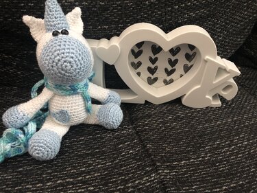 Weißes und hellblaues gehäkeltes Einhorn-Amigurumi mit blau-türkisem Schal neben einem weißen Herzrahmen.
