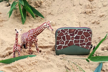 Kleine Reißverschluss-Geldbörse mit Giraffenmuster und grünem Boden im Sand neben zwei Giraffenfiguren