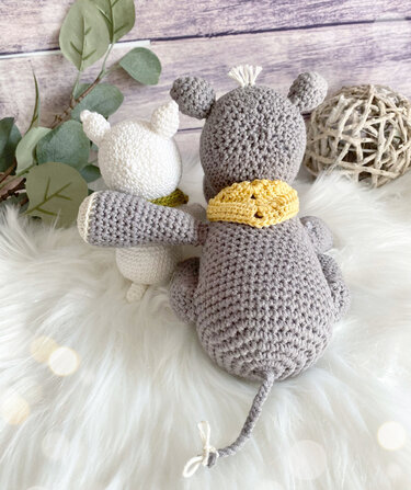 Rückansicht: zwei gehäkelte Amigurumi‑Mäuse, eine grau mit gelbem Schal, eine weiß mit grünem Schal, auf weichem weißem Untergrund; dezente Deko im Hintergrund.