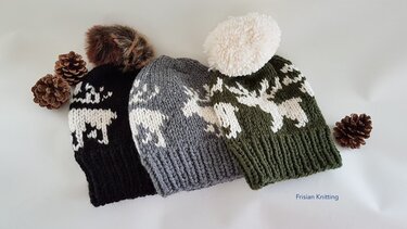 Drei grob gestrickte Beanies in Schwarz, Grau und Olivgrün mit weißem Rentiermotiv und unterschiedlichen Bommeln, auf hellem Hintergrund neben Tannenzapfen.