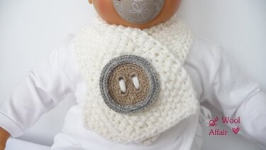 Cremeweißer gehäkelter Babyschal mit großem runden Zierknopf in Braun und Grau, an einem Baby getragen.