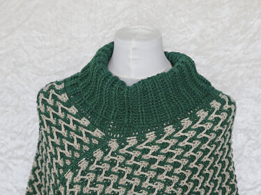 Gehäkelter Damen-Poncho in Dunkelgrün und Beige mit grafischem Zickzackmuster und hohem Rollkragen auf einer Schneiderpuppe.