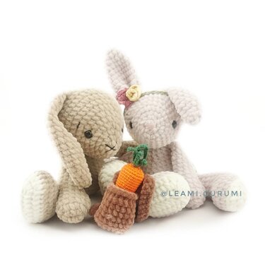 Zwei flauschige gehäkelte Amigurumi-Hasen (beige und hellrosa) mit Blumenstirnband und einer kleinen gehäkelten Karotte.