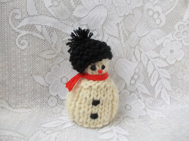 Gestrickter Mini‑Schneemann in Creme mit schwarzer Bommelmütze und rotem Schal vor Spitzenstoff