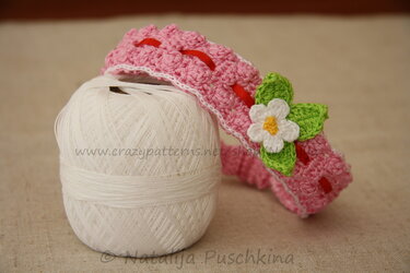 Rosa gehäkeltes Baby-Stirnband mit weißer Häkelblume, grünen Blättern und rotem Band auf weißem Garnknäuel