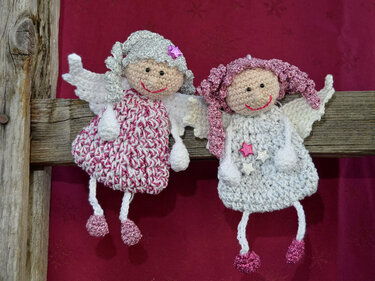 Zwei gehäkelte Engelchen in Rosa und Weiß mit Flügeln und glitzernden Haaren an einer Holzleiste vor rotem Hintergrund