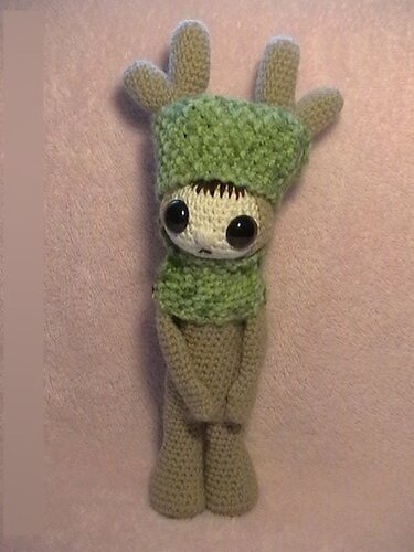 Handgehäkeltes Amigurumi mit hellbraunem Körper, grüner Mütze mit geweihartigen Spitzen, grünem Schal und großen dunklen Augen auf hellem Hintergrund.