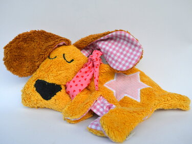 Liegender gelb-oranger Kuscheltier-Hund mit rosa-kariertem Innenohr, Herzchen-Halstuch und Stern-Applikation vor hellem Hintergrund
