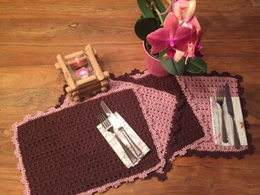 Gehäkelte rechteckige Tischsets in Braun und Rosa auf Holztisch mit Besteck, Kerze und Orchidee