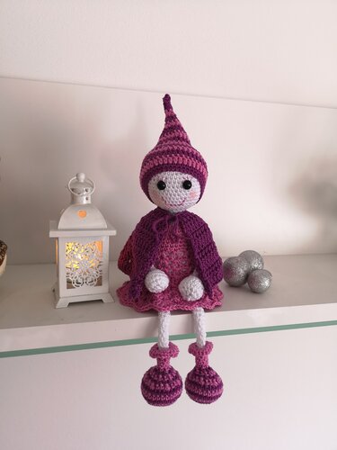 Handgehäkelte Amigurumi-Puppe in Lila und Rosa mit gestreifter Zipfelmütze auf weißem Regal neben einer Laterne