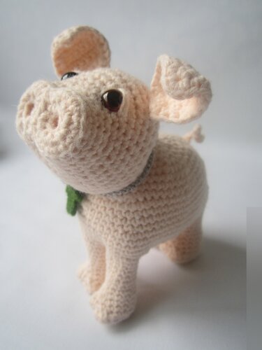 Helles gehäkeltes Amigurumi-Schwein mit aufgerichtetem Kopf, modellierter Schnauze und kleinem grünen Kleeblatt am Hals auf weißem Hintergrund.