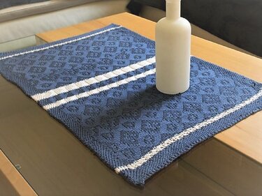 Blau-weißer gestrickter Tischläufer mit Rautenmuster und zwei weißen Querstreifen auf einem Glas- und Holztisch, mit weißer Vase darauf