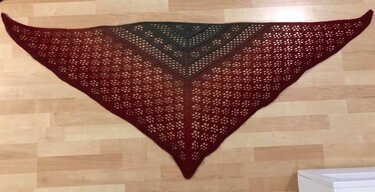 Crocheted triangular ombre shawl in burgundy to charcoal grey with a repeating lace motif laid flat on a wooden floor.
