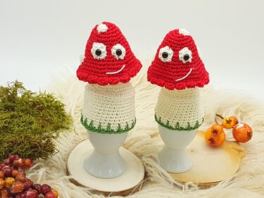 Zwei rote, gehäkelte Fliegenpilz-Eierwärmer mit weißen Punkten auf Eierbechern, davor Moos und Deko-Beeren