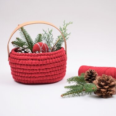 Round red crocheted basket with tan handle filled with evergreen sprigs, pine cones, and a red crocheted ornament; red yarn and extra pine cones nearby.