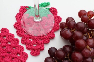 Gehäkelter pinker Untersetzer in Beerenform mit grünem Blatt, Weinglas darauf, rote Trauben daneben