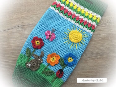 Gehäkeltes Utensilo in Blau und Grün mit applizierten Motiven: Sonne, Schmetterling, drei Blumen, Hase und Marienkäfer.