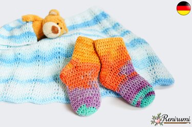 Handgehäkelte Babysocken in Orange, Lila und Türkis auf blau-weißer Häkeldecke mit kleinem Teddybär