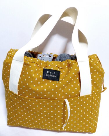 Senfgelbe gepunktete Stofftasche mit weißen Henkeln, gef		llt mit Strickutensilien und einem Ma		band in der Au		entasche.