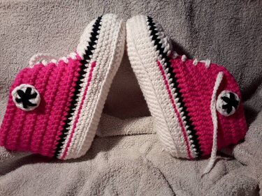 Zwei pink-weiße gehäkelte Hausschuhe im Sneaker-Stil mit Schnürsenkeln auf grauem Handtuch.