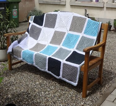 Gehäkelte Decke aus Granny Squares in Grau, Türkis und Weiß auf einer Holzbank