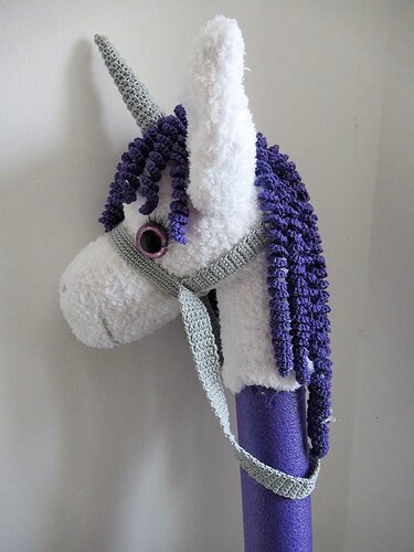 Gehäkeltes weißes Einhorn-Steckenpferd mit lila Mähne, grauem Horn und Zaumzeug auf lila Stab