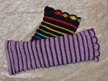 Zwei gehäkelte Armstulpen auf hellem Untergrund: lila Rippenstulpen und schwarze Stulpen mit bunten Regenbogenstreifen.