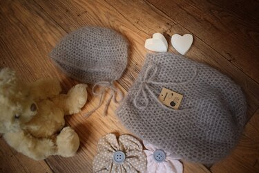 Flauschiger gehäkelter Babykokon mit Mütze, liegend neben Stoffblumen, Holzherzen und Teddybär