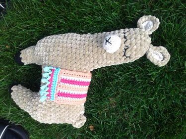 Beiges gehäkeltes Amigurumi-Tier mit gestreiftem Sattel, liegend auf Gras, geschlossene Augen gestickt