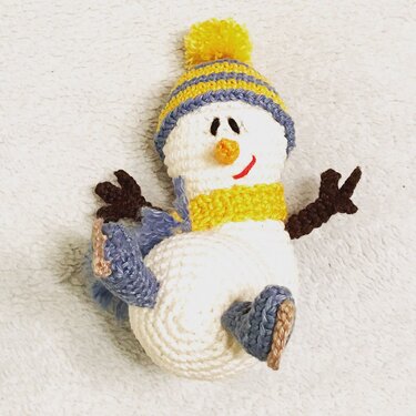 Gehäkelter Amigurumi-Schneemann mit gelb-blau gestreifter Mütze, gelbem Schal, braunen Armen und blauen Fäustlingen auf hellem Hintergrund.
